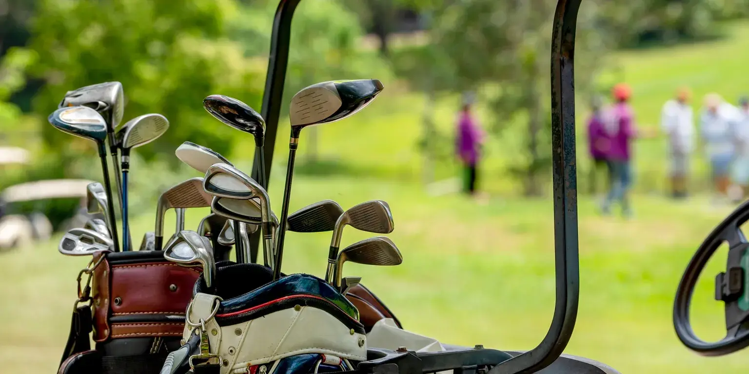 Golfbagar med klubbor på en golfbil, med spelare på banan i bakgrunden