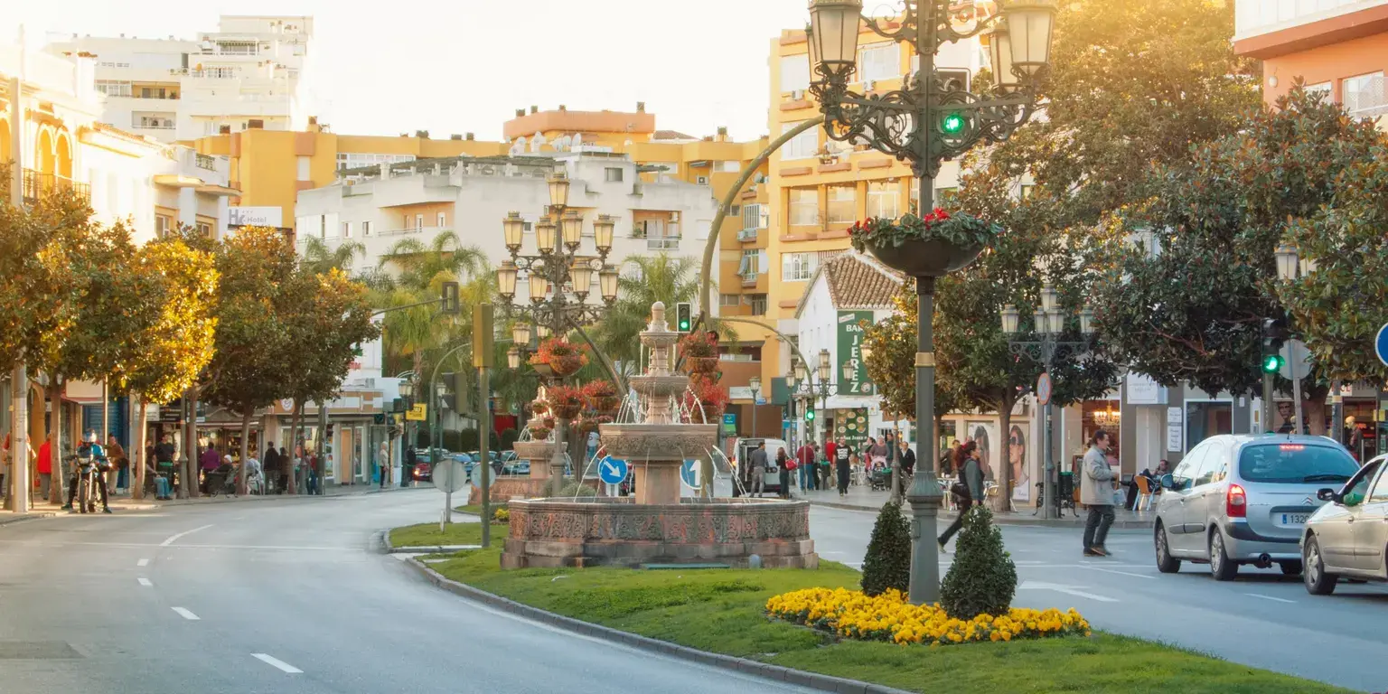 Gata i Torremolinos med fontän, blomsterrabatt och gula bostadshus i kvällssol